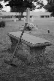 A rusted pickaxe at the entrance to a grave yard, leaning on a stone bench.